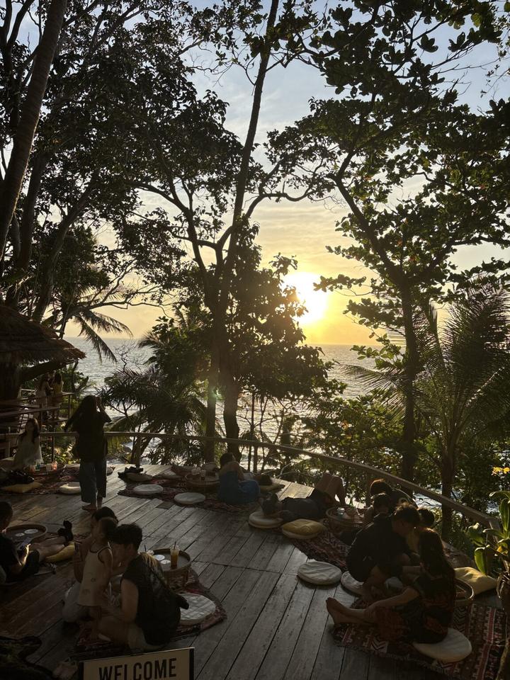 Sunset over the sea, seen through the trees from a terrace where guests sit and watch
